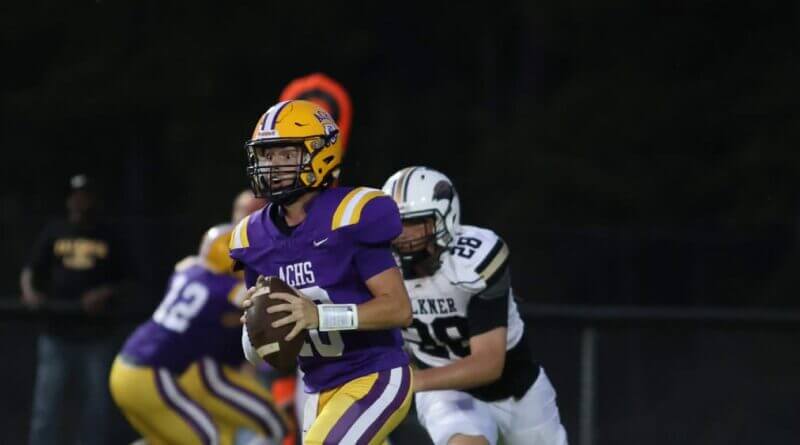 An Alcorn Central football player in a purple jersey and gold helmet holds the ball as a defender approaches.